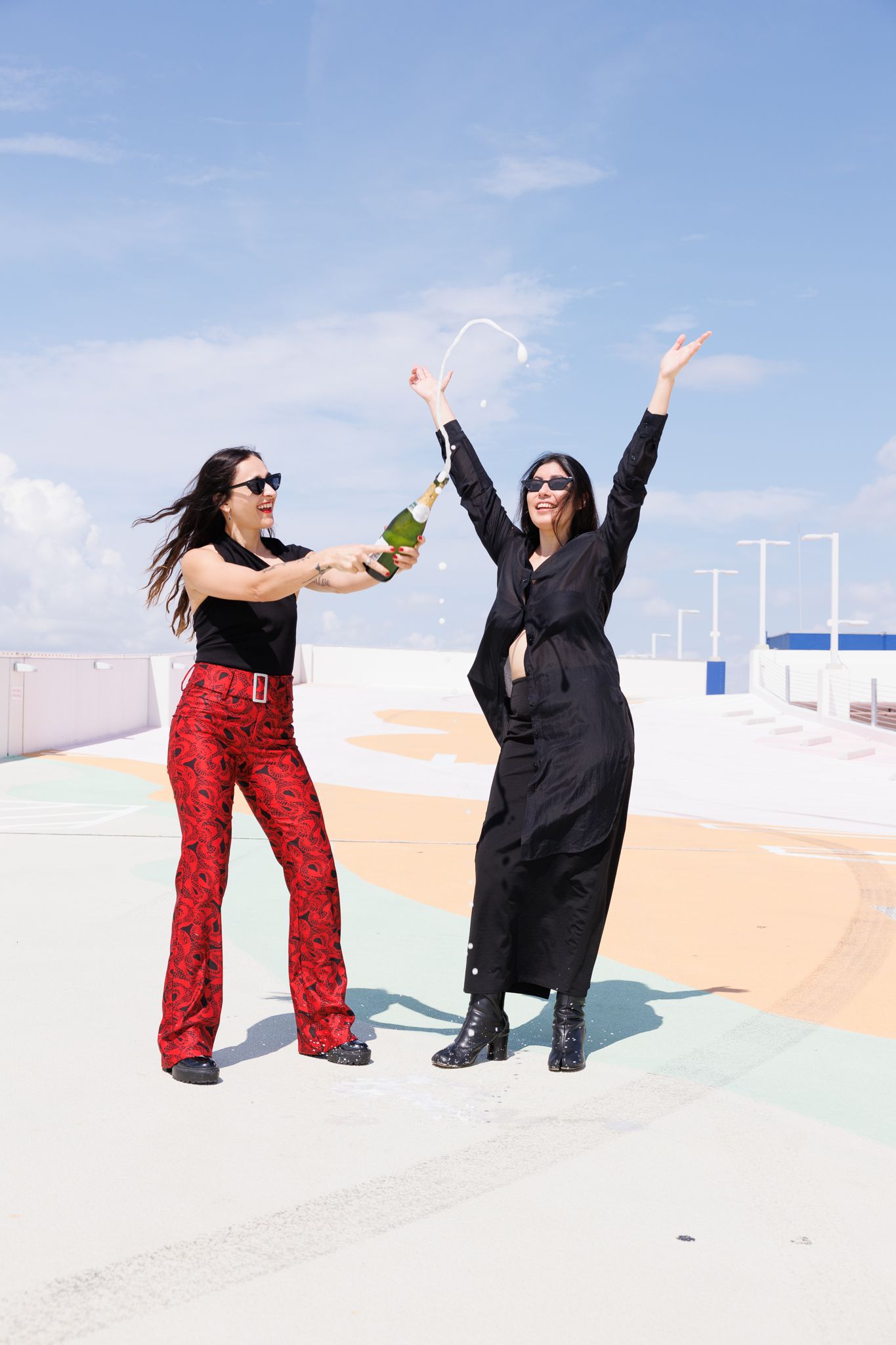Two friends celebrating on a rooftop, one wearing red pants and another in black, jumping with joy against a blue sky.