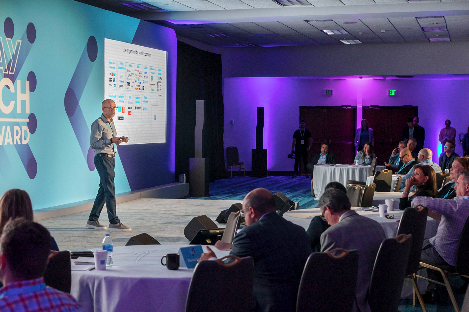 Speaker presents at tech conference with projection screen and seated audience in dimly lit room for an Event Photography in Miami