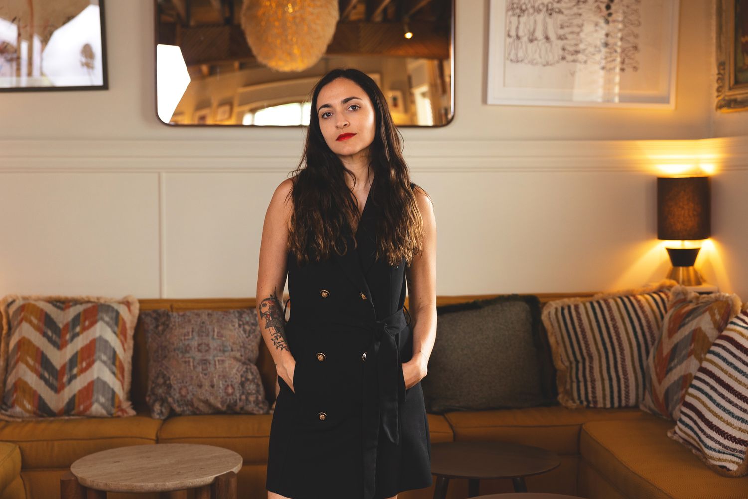 A person in a black sleeveless dress stands confidently in a warmly lit restaurant with lounge seating.