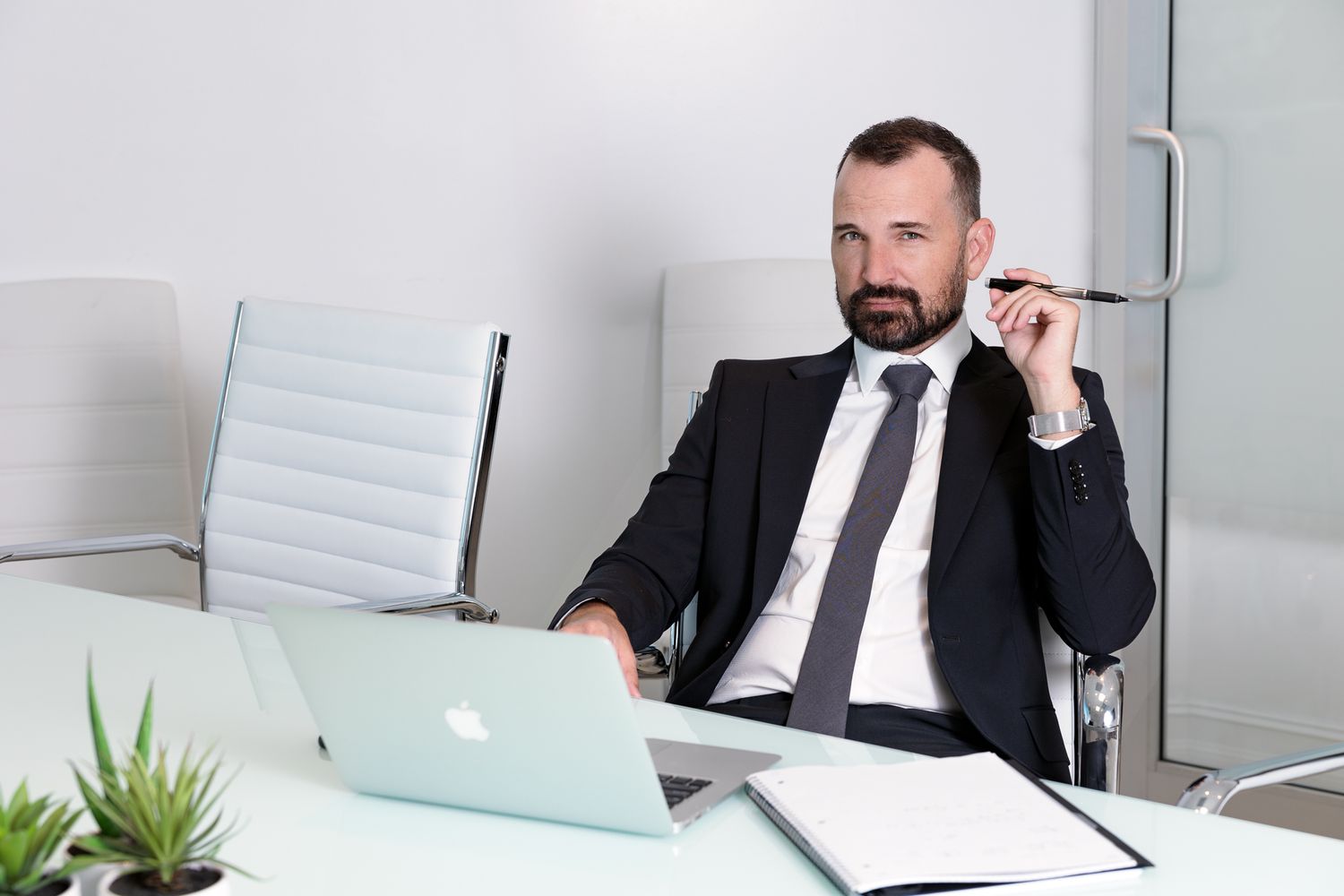 Professional man in dark suit sitting at modern desk with laptop in minimalist office setting in a personal branding photography shoot