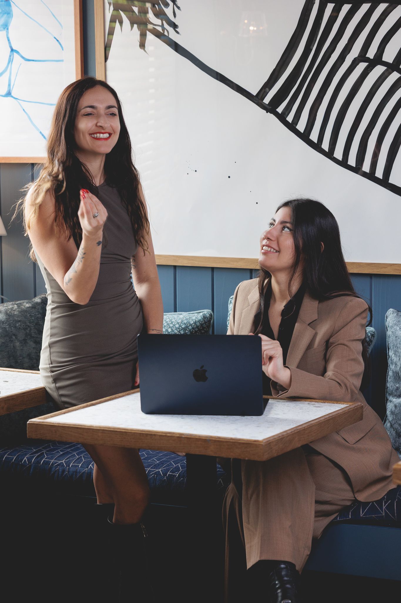 Two colleagues share a lighthearted moment while working together with a MacBook laptop at a cafe table.