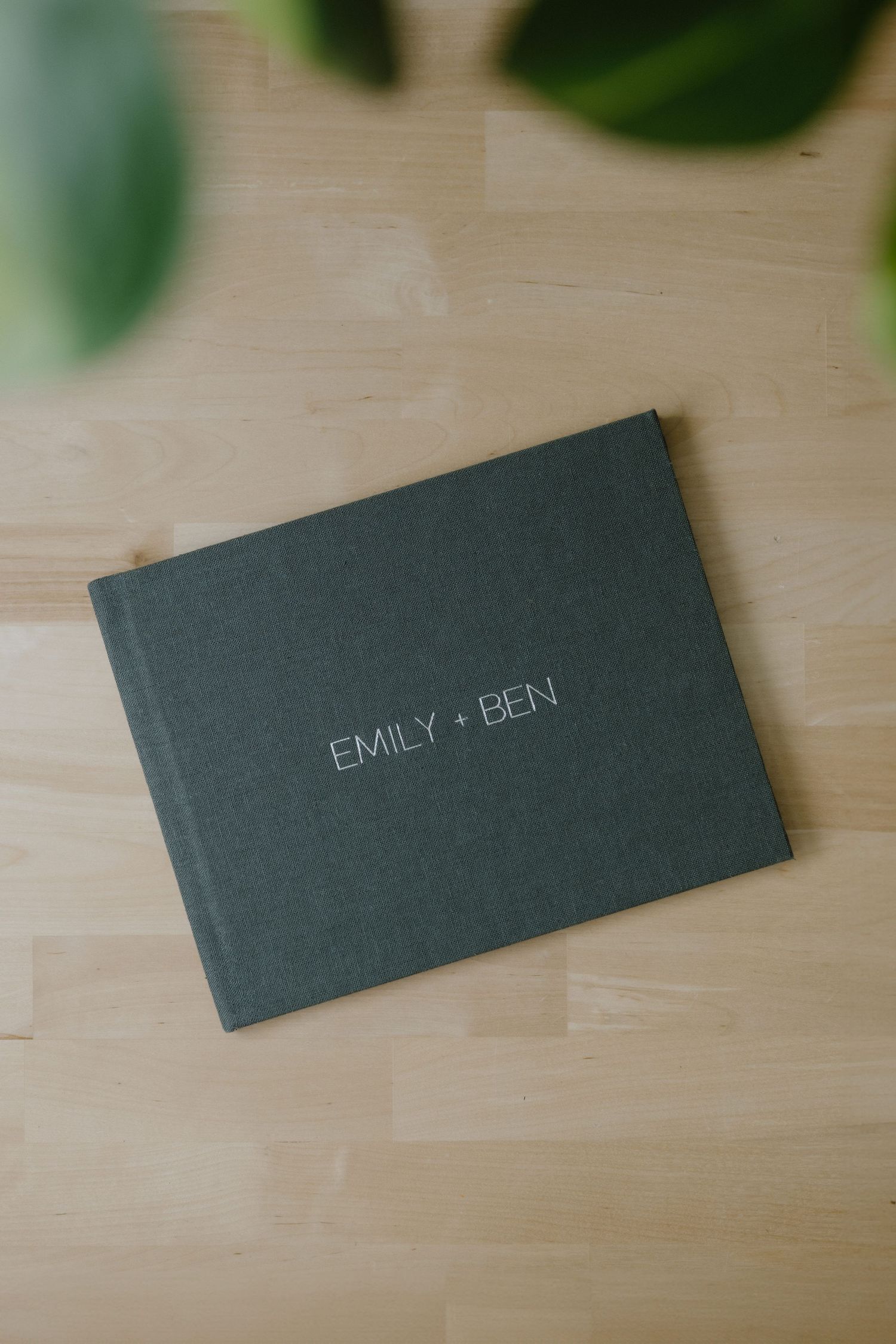 A grey album book with 'EMILY + BEN' text on the cover sits on a wooden surface with green leaves visible in background.