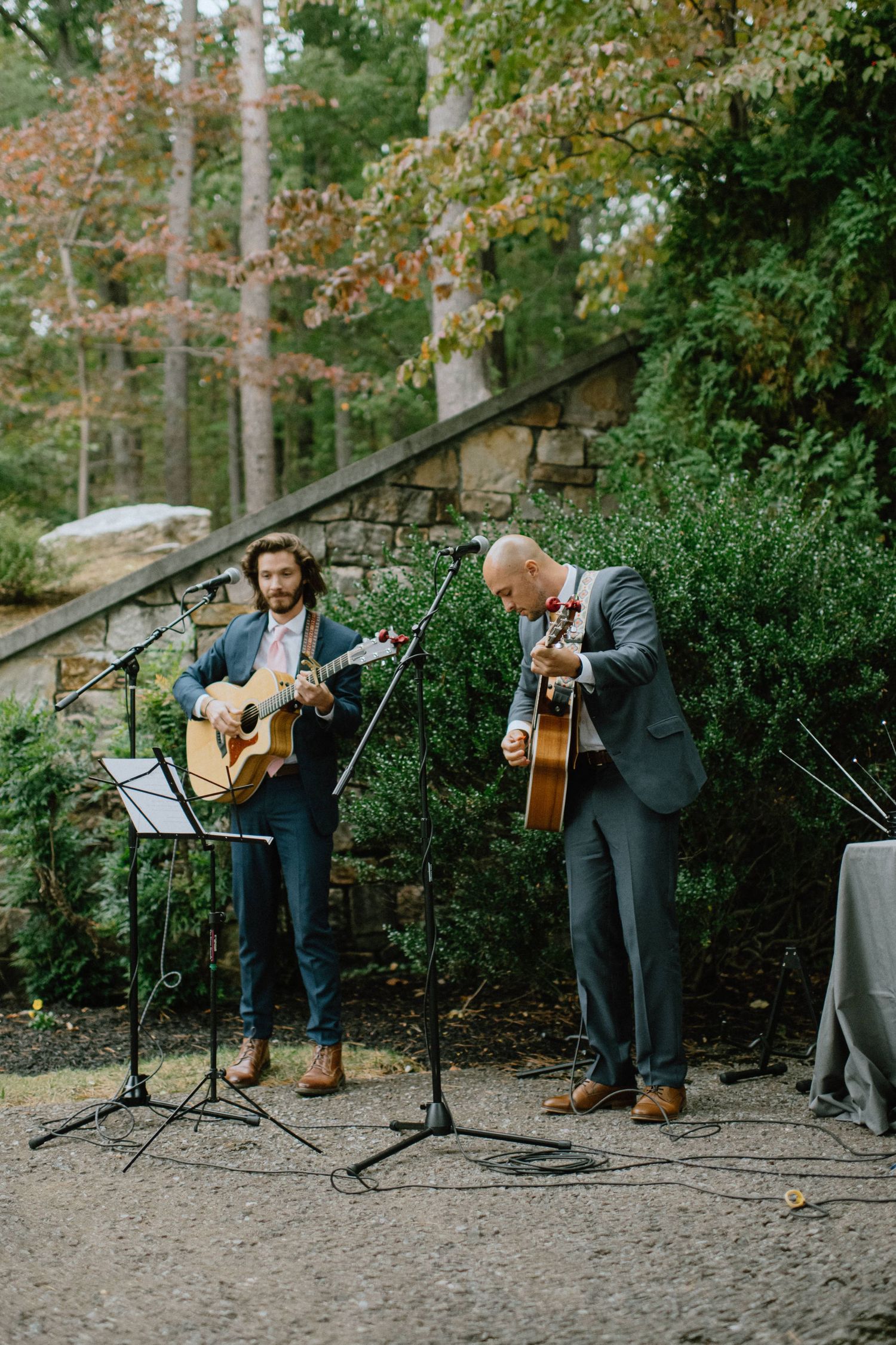 Two musicians performing with acoustic guitars outdoors against a stone wall backdrop.