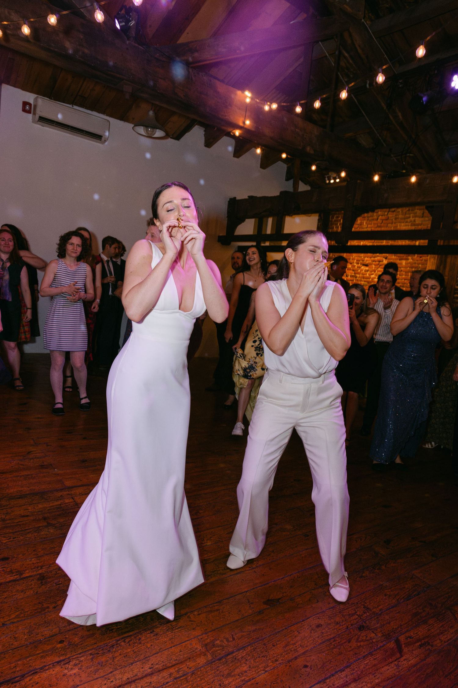 Two people in white outfits showing joyful reactions during a wedding reception with string lights and purple ambiance.