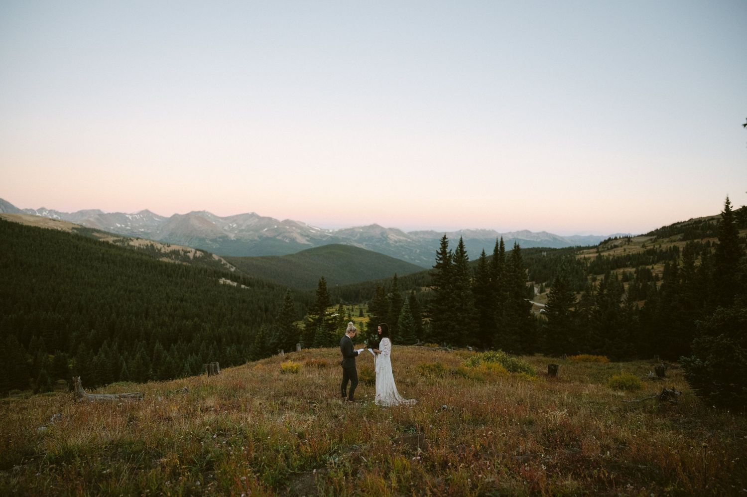Couple standing on mountain overlook during sunset with expansive mountain range views.