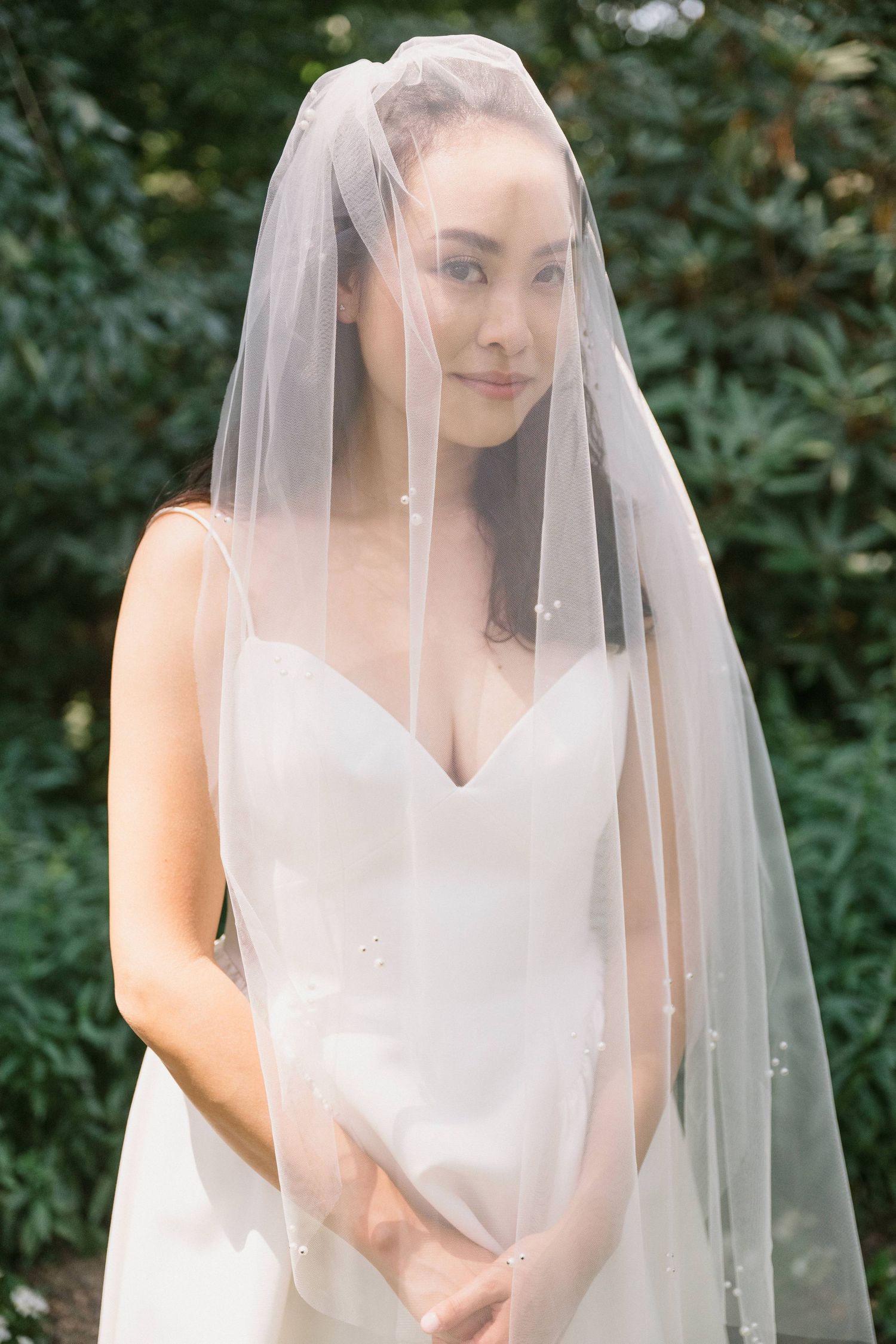 Bride in white wedding dress poses delicately under sheer veil in garden setting.