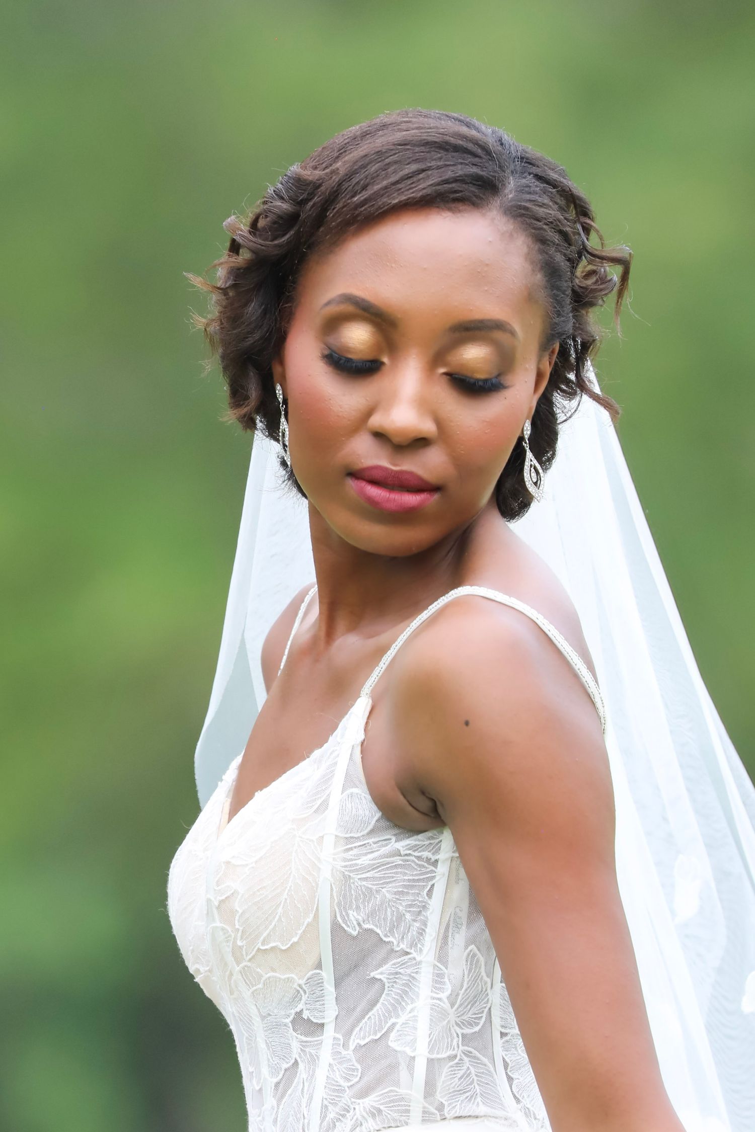 A bride in a white lace wedding dress with gold eye makeup and a flowing veil poses with closed eyes against a green background.