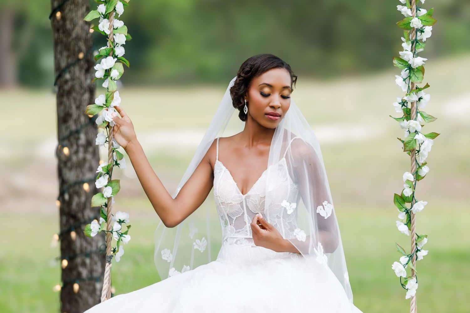 A bride in a flowing white wedding dress poses on a floral swing in an outdoor garden setting.