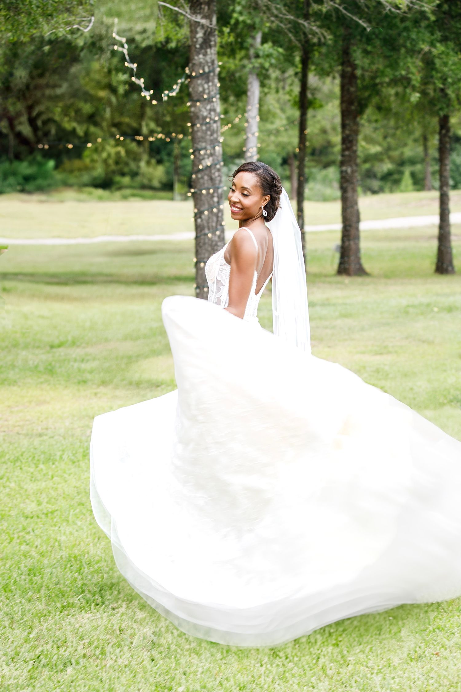 A bride in a flowing white wedding dress twirls joyfully on a grassy lawn surrounded by trees on a sunny day.