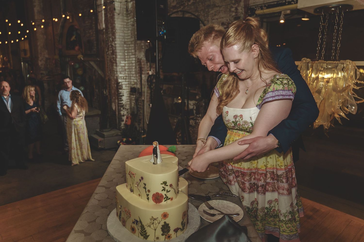 A couple cuts their rustic wedding cake decorated with greenery and flowers in a warm-lit reception venue.
