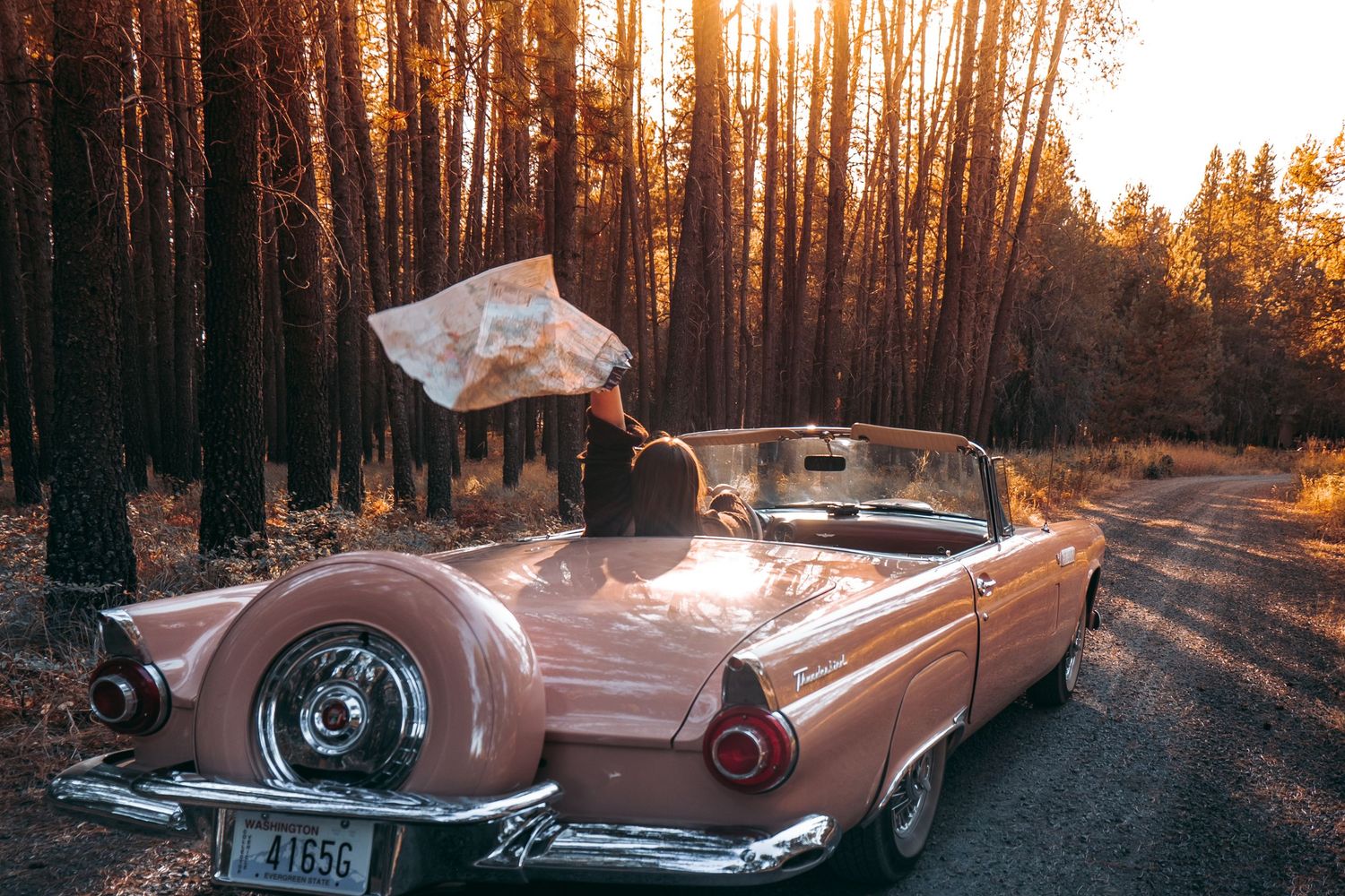 Pink vintage convertible car parked on forest road at sunset with map floating in golden light.