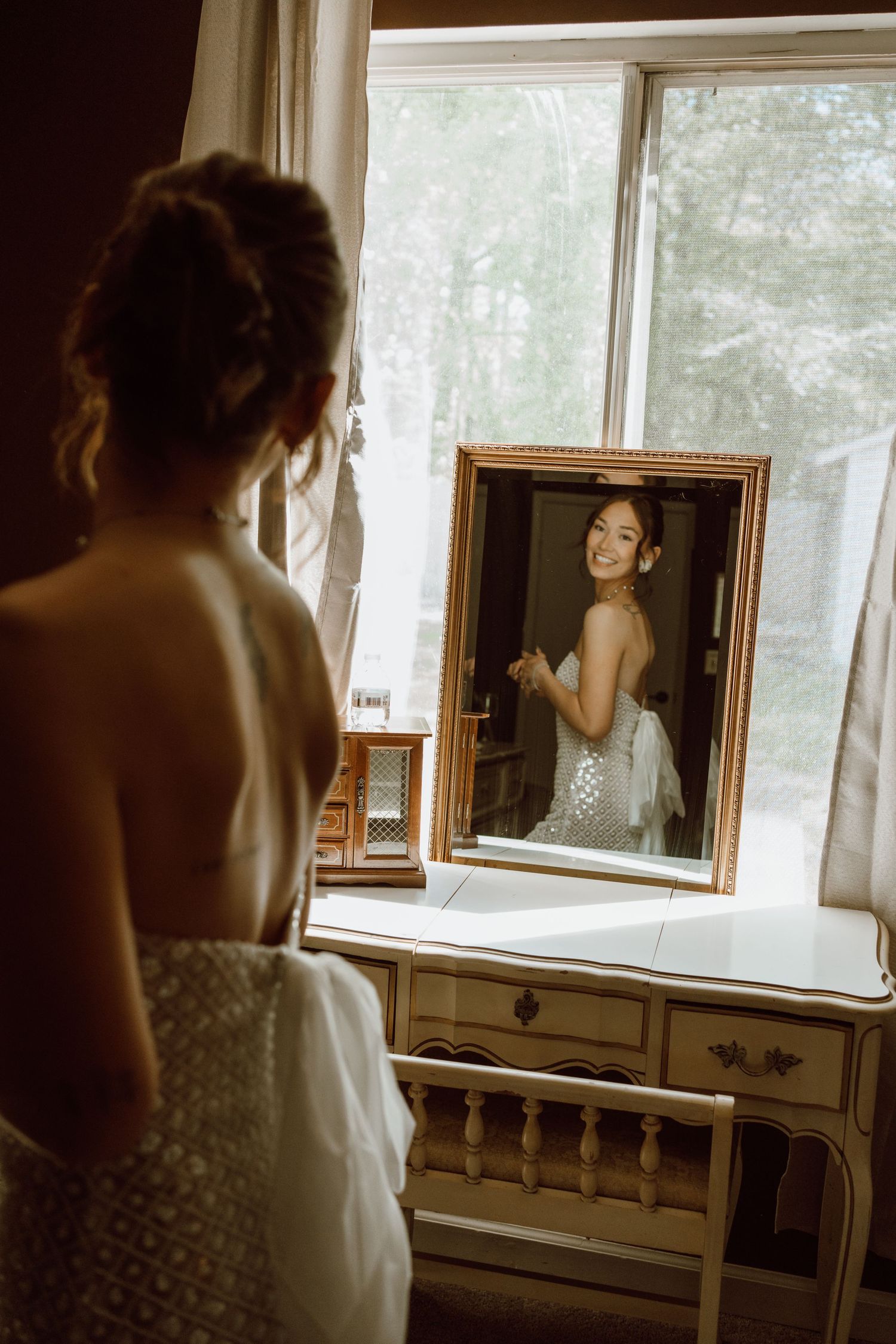 A person wearing a white dress looks at their reflection in an antique vanity mirror by a window.