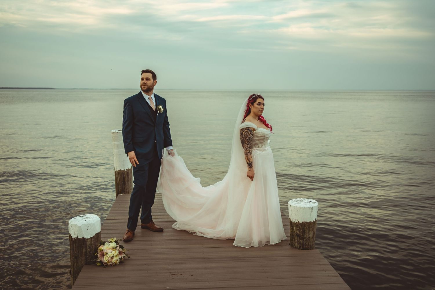 A couple stands on a wooden dock over water at sunset, with the bride's white dress flowing in the breeze.