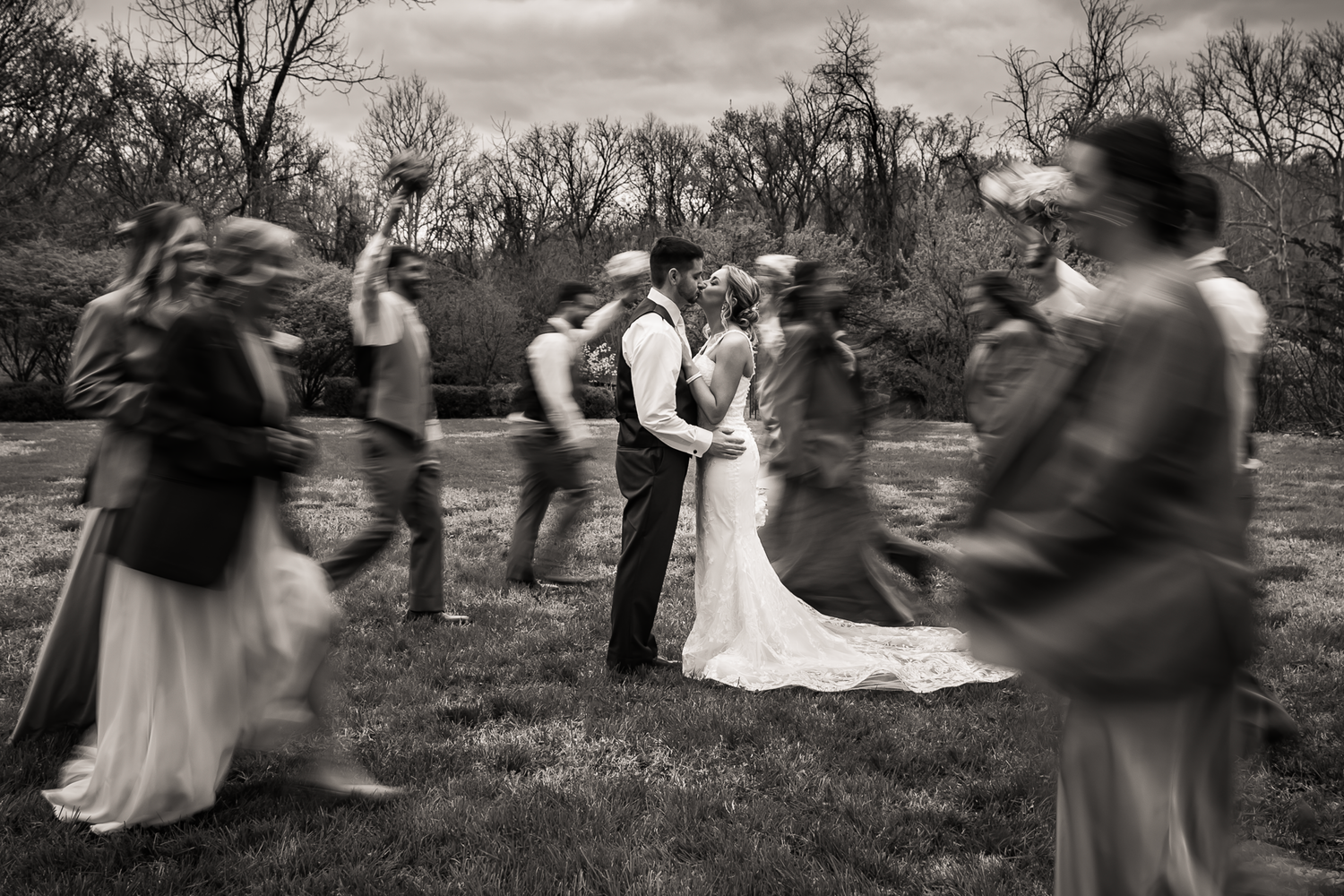 A black and white photograph of outdoor dancing couples in motion during a rustic wedding celebration in nature.