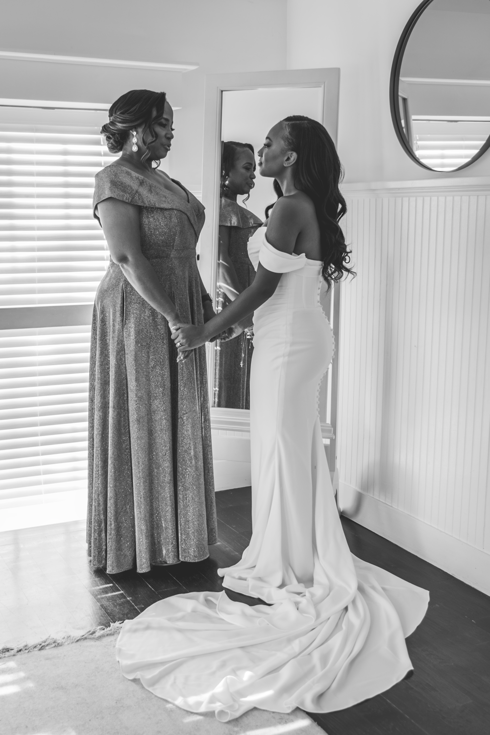A tender black and white moment between a bride in a flowing white gown and someone in a lace dress by a window.