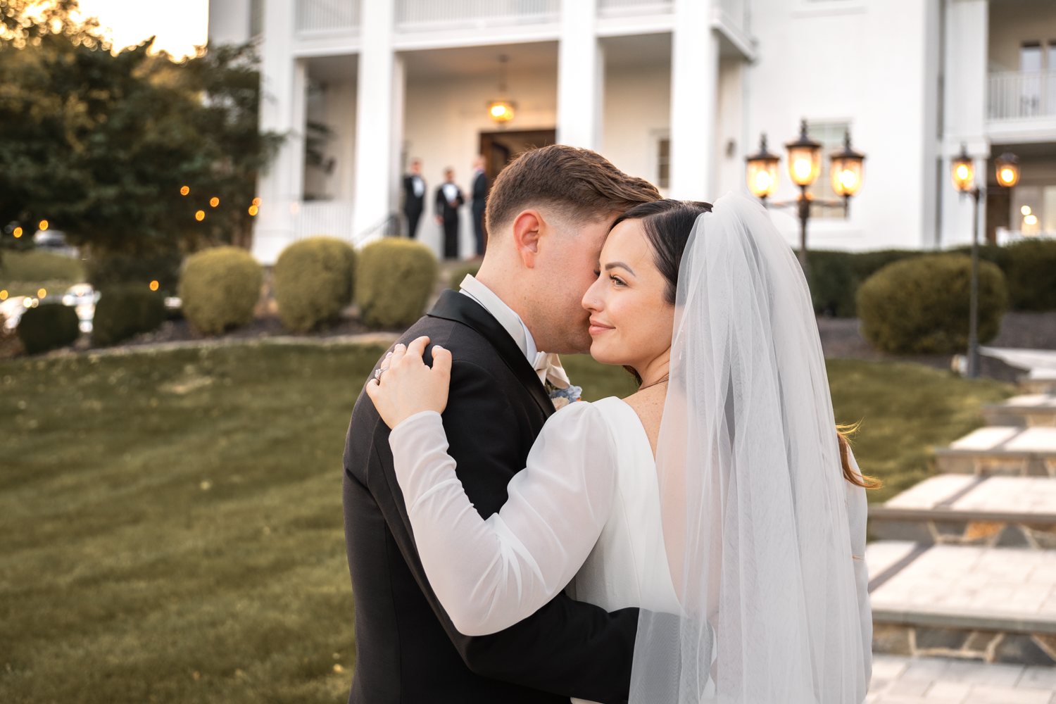 Bride and groom share an intimate embrace during sunset outside an elegant white columned venue.