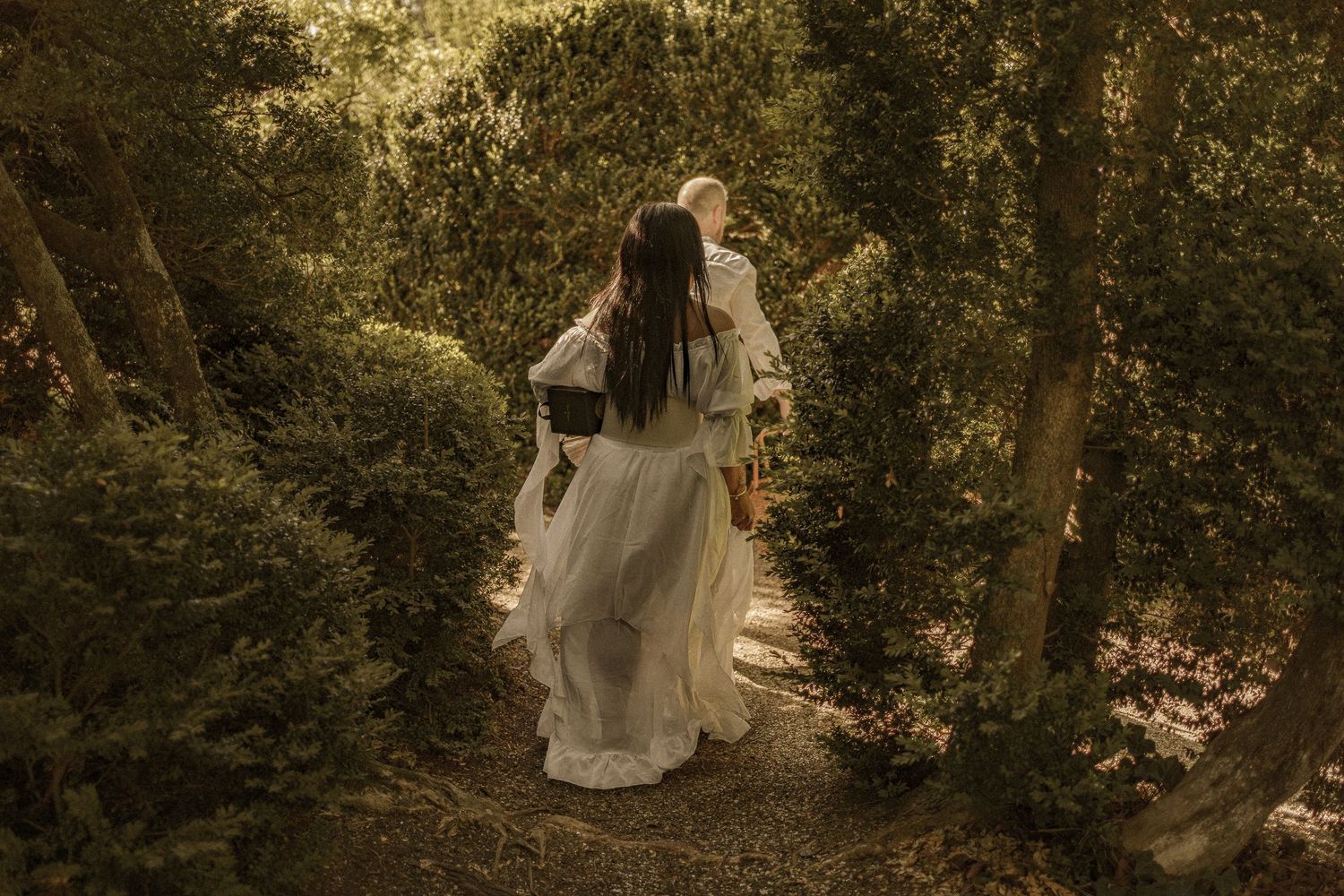A romantic scene in the forest with figures in flowing white clothing walking down a wooded path at golden hour.