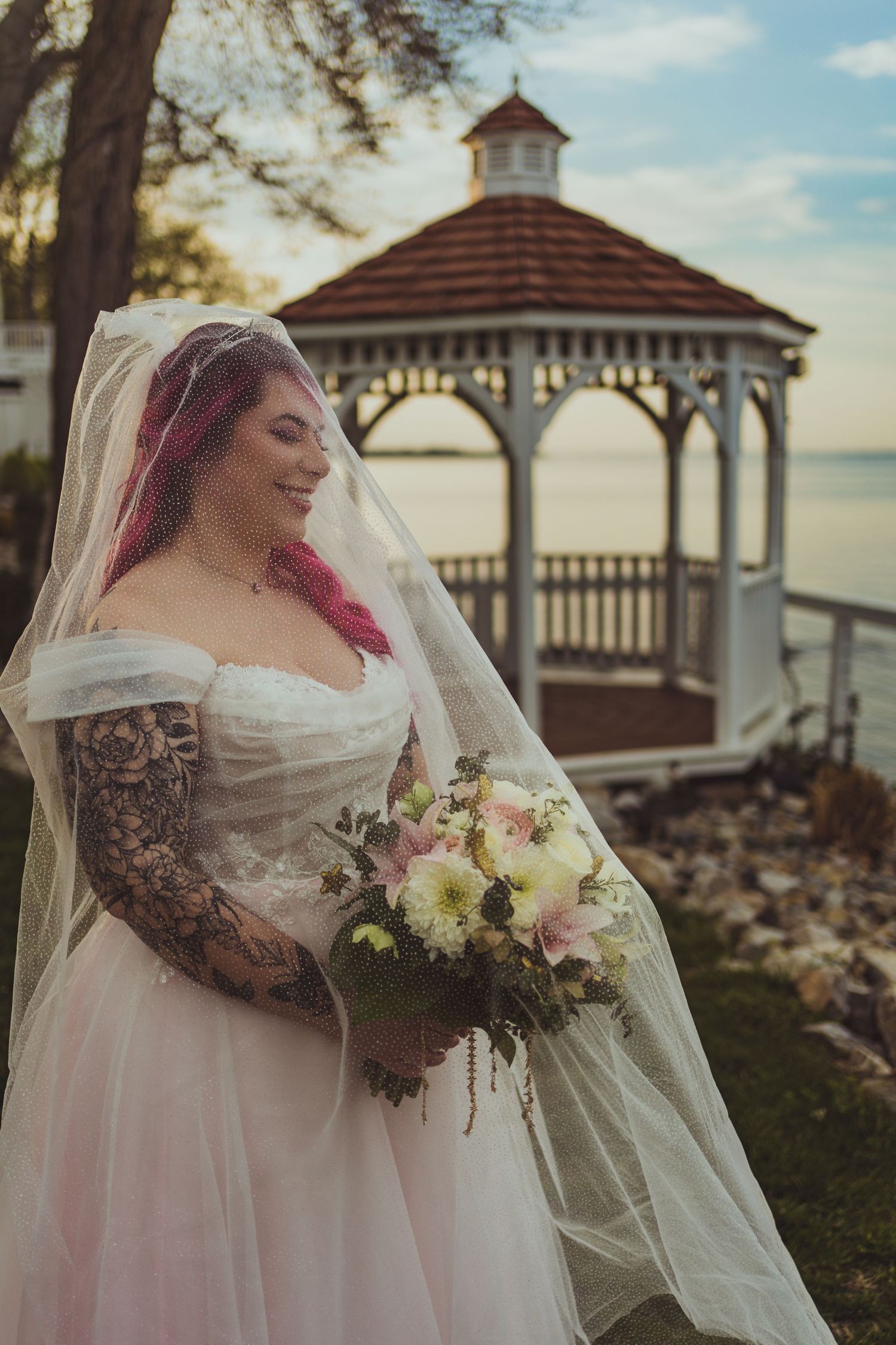 A bride in a white dress with tattoos holds a pastel bouquet while standing by a waterfront gazebo at sunset.