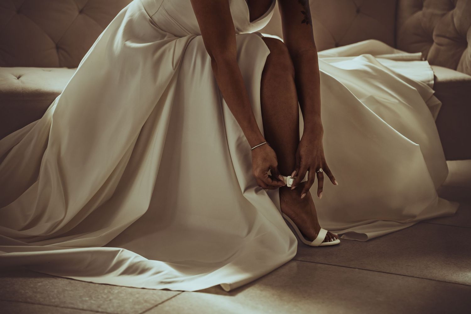 Person wearing flowing white wedding dress adjusts elegant heels while seated on brown leather couch.