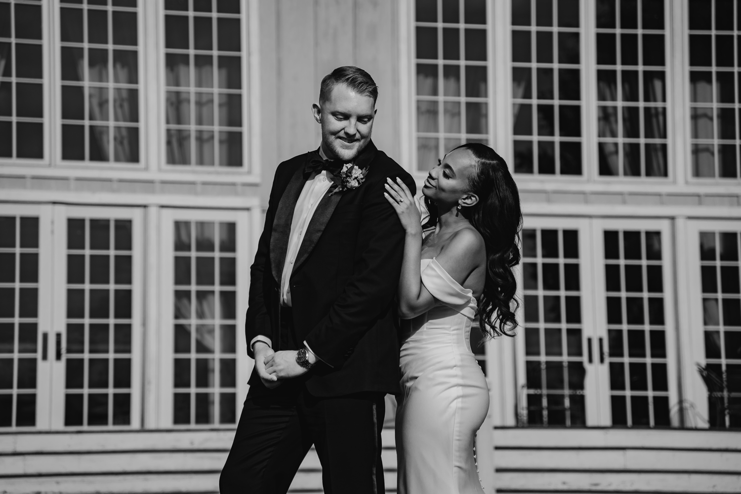 A couple in wedding attire share a candid moment in black and white in front of large glass windows.