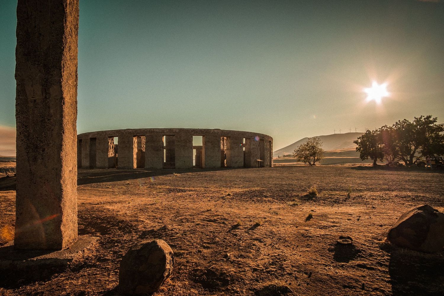 A replica of Stonehenge stands in silhouette against a bright sun and dusty landscape at sunset.