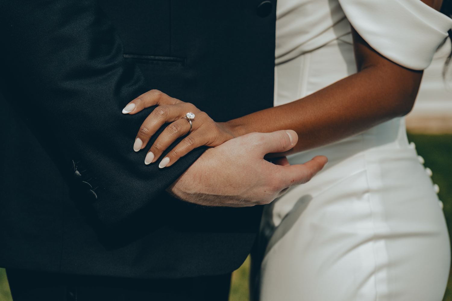 Close-up of hands embracing during wedding ceremony, with black suit and white dress visible.