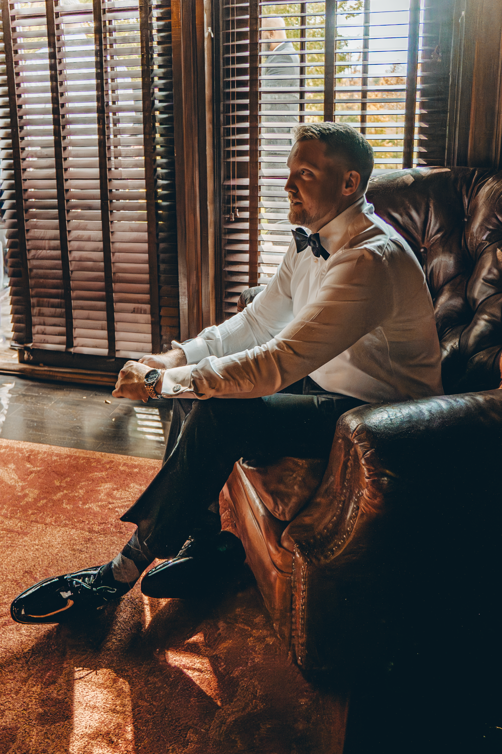 A person sits in a leather armchair by wooden shuttered windows wearing a white dress shirt and dark pants.