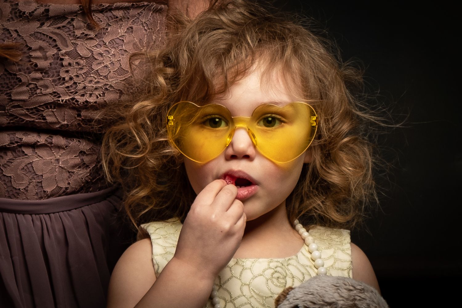 Child wearing yellow-tinted glasses and lace dress strikes contemplative pose against dark background.
