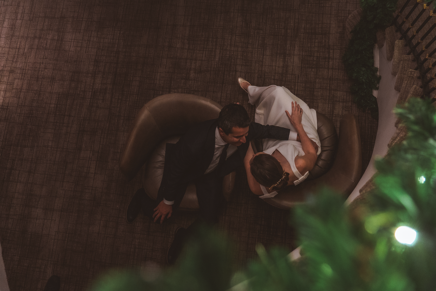 A person wearing business attire bends down to examine something through lush green foliage in low light.