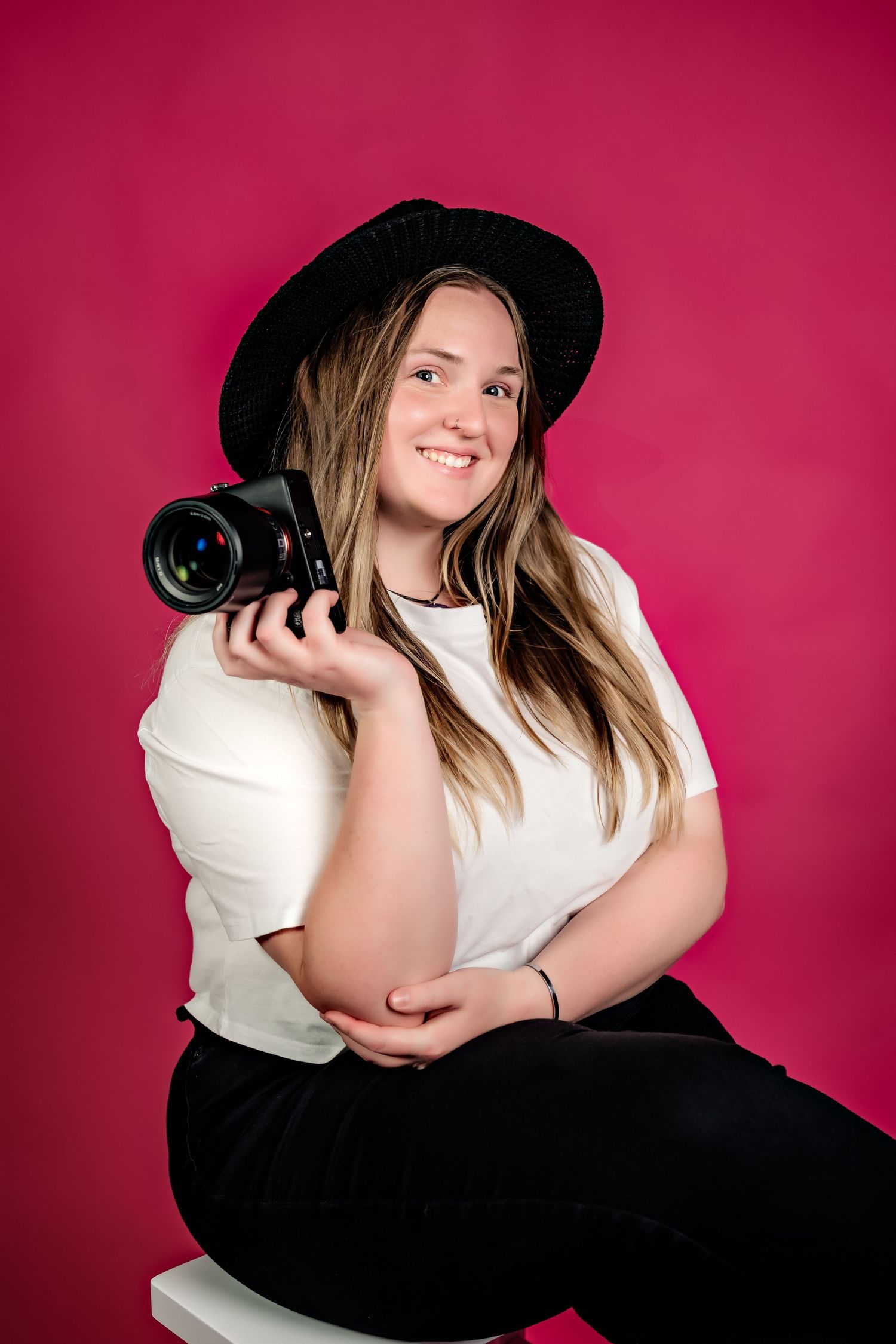 Portrait of photographer wearing black hat and white top holding camera against pink background.