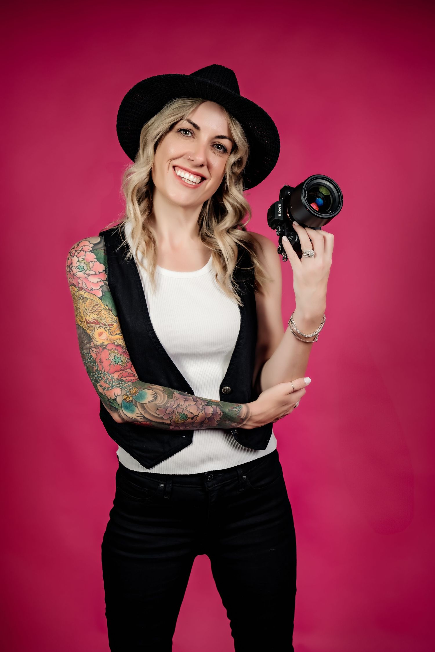 Portrait of photographer with tattoo sleeve wearing black hat and vest holding camera against pink background.