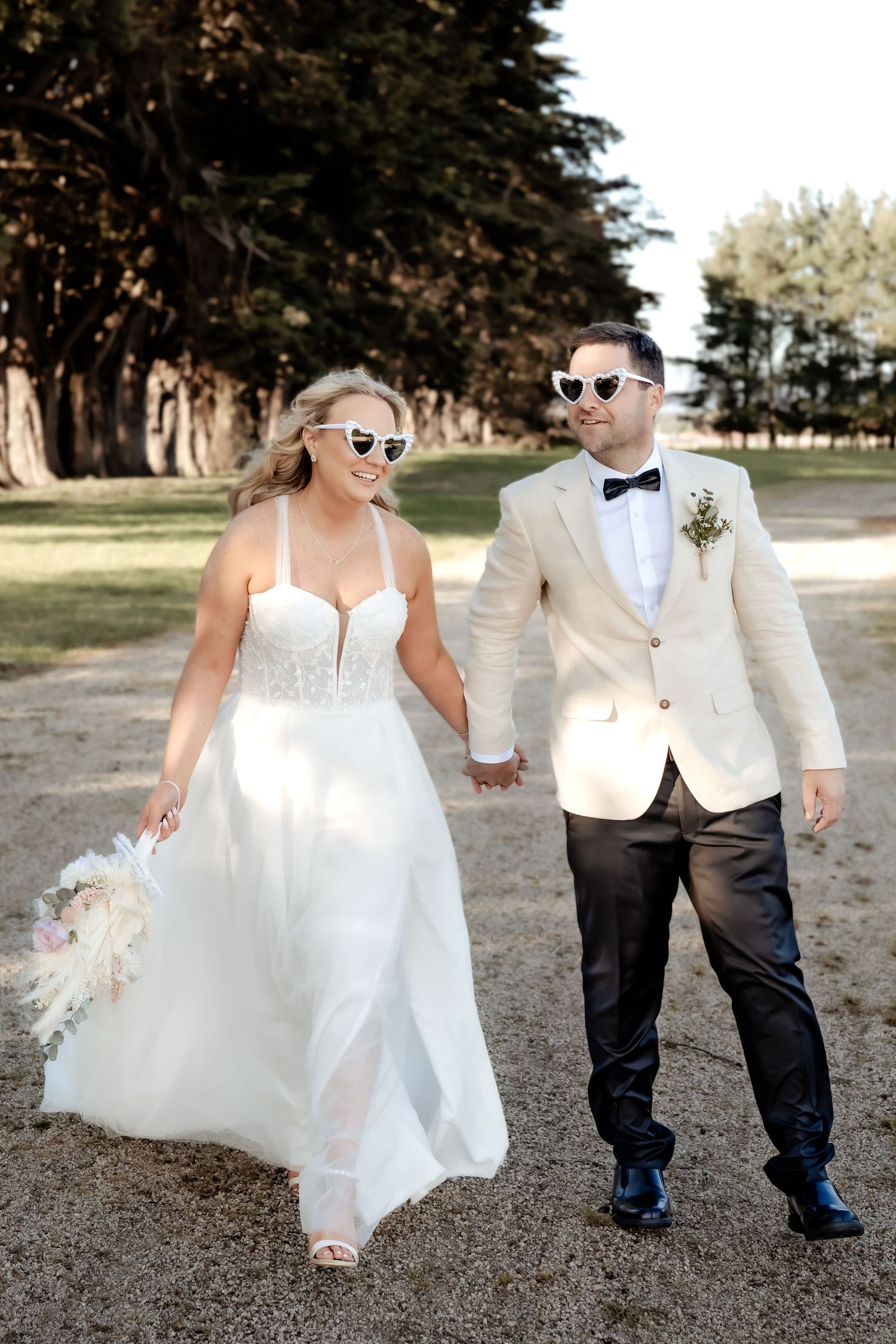 Couple wearing matching sunglasses walking hand in hand at their outdoor wedding with white dress and light colored suit.