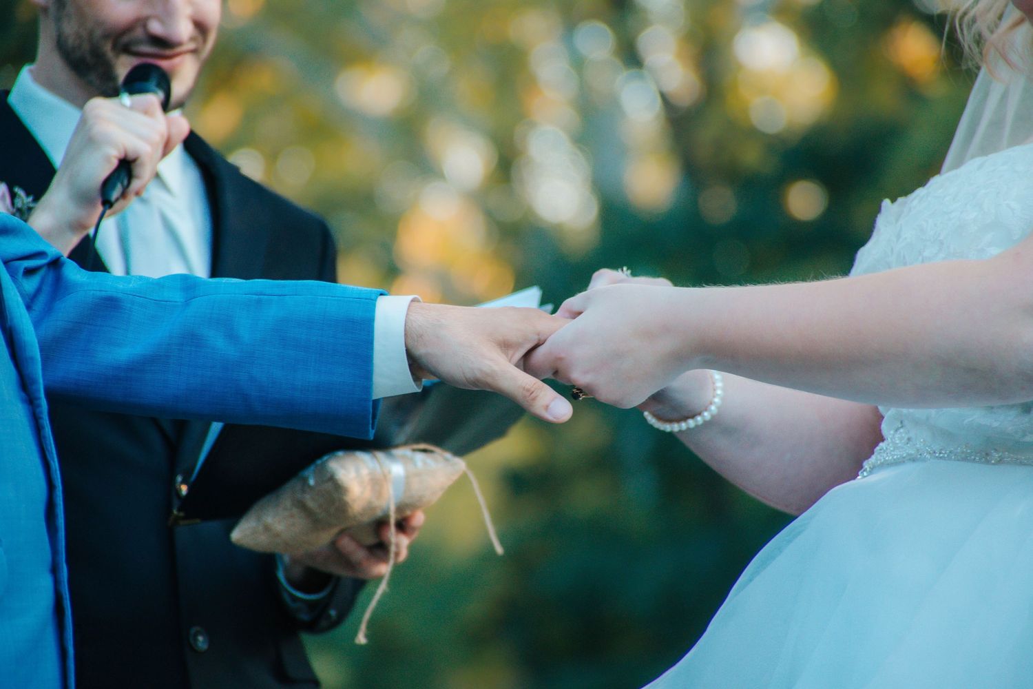 A romantic wedding ceremony moment showing hands exchanging rings against a soft sunlit outdoor backdrop.
