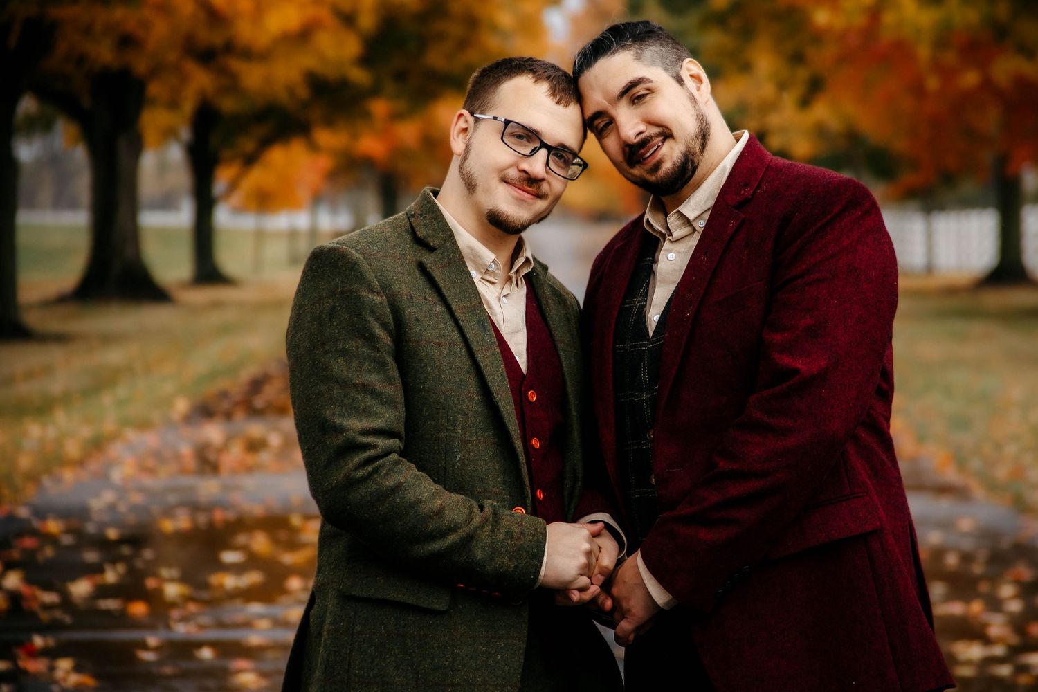 Two people wearing wool coats stand close together on an autumn day with orange fall foliage in the background.