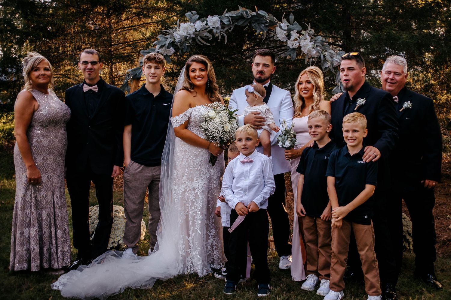 Wedding party group stands together outdoors with trees in background during golden hour.