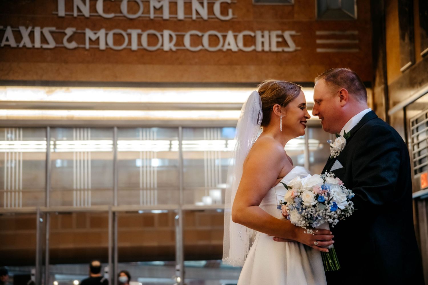 Newlywed couple shares intimate moment under vintage bus station signage while holding blue and white wedding bouquet.