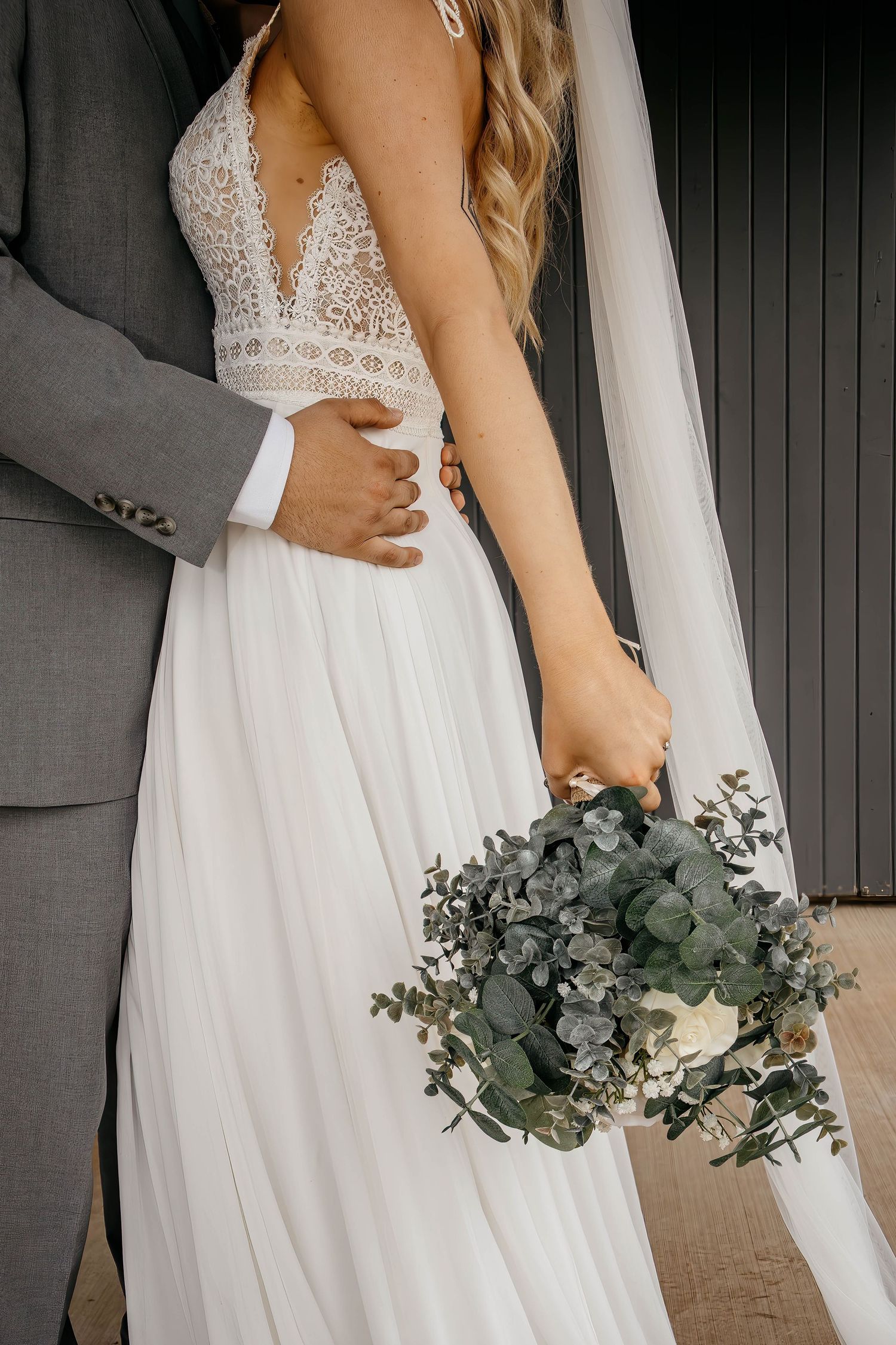 Bride in lace v-neck wedding gown with ethereal eucalyptus bouquet embraced by groom in grey suit against dark wall.