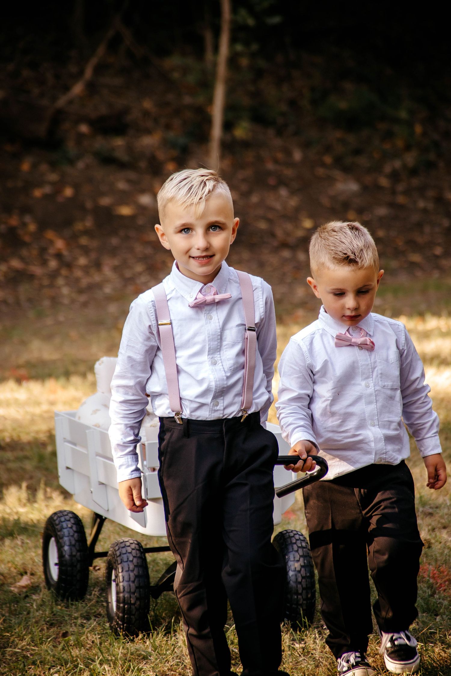 Two young boys in matching white shirts, bow ties and suspenders stand next to a rustic wooden wagon outdoors.