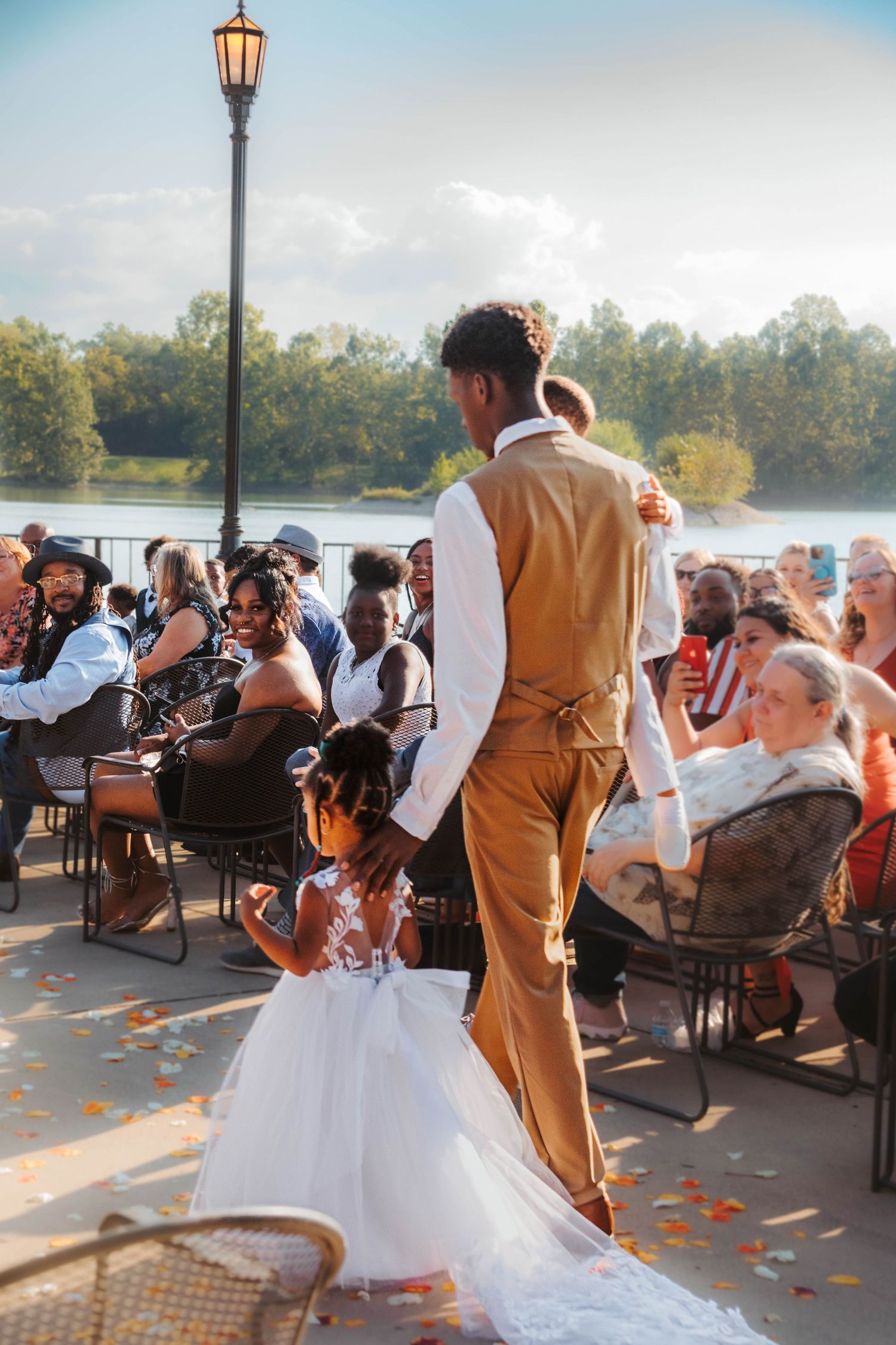 A wedding reception outdoor by a lake with guests seated at tables while autumn leaves scatter across the patio floor.