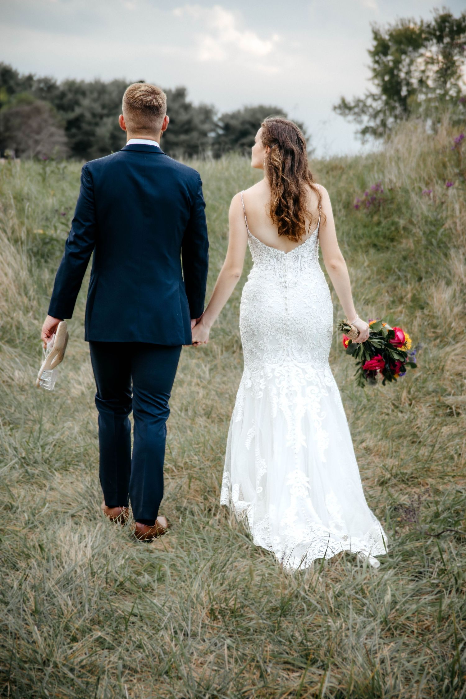 Couple walks through meadow on wedding day with bride in lace gown holding red bouquet and groom in navy suit.