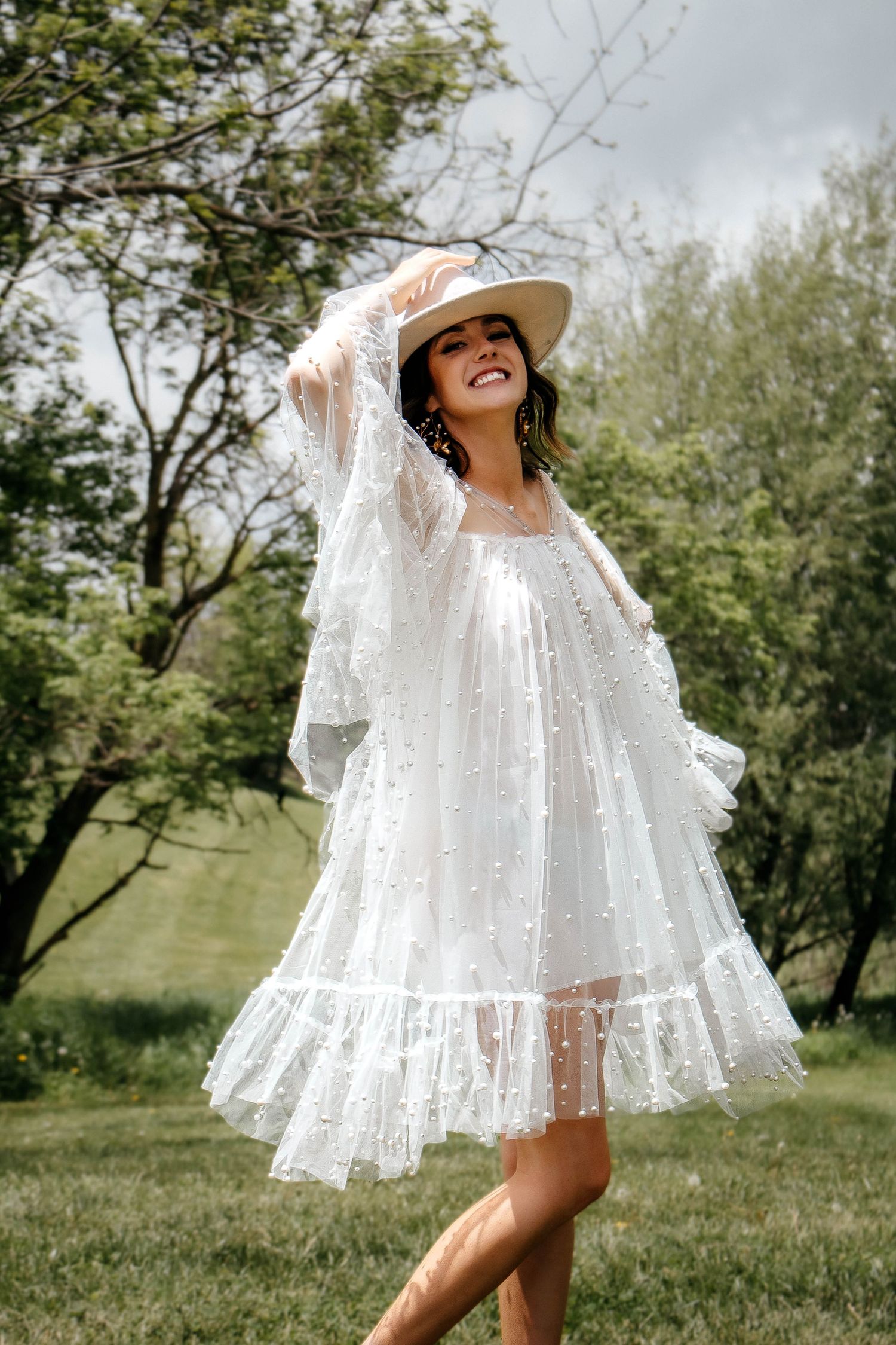 A joyful summer moment captured outdoors featuring a flowing white lace dress and straw sunhat against a natural backdrop.