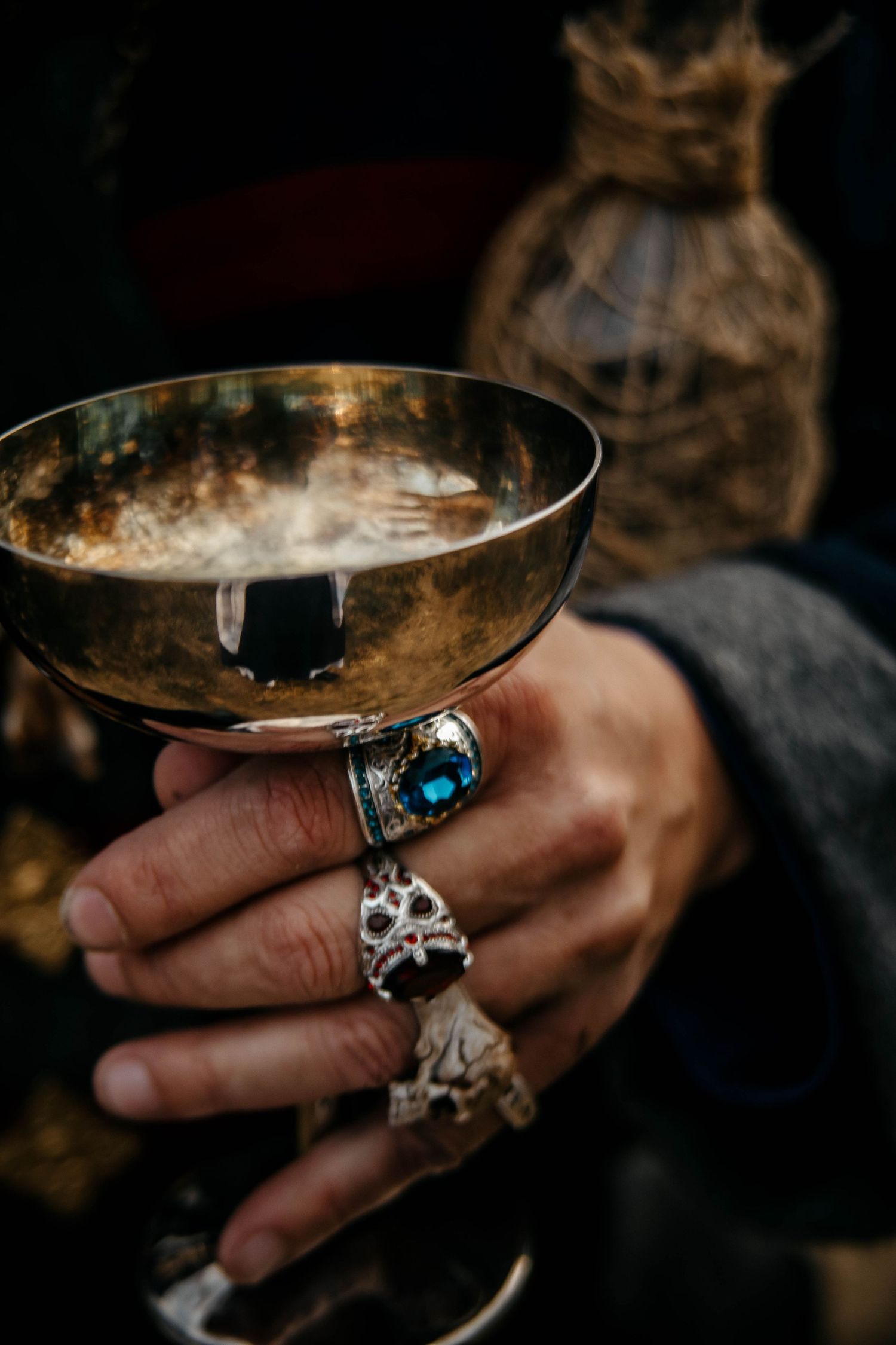 A hand wearing ornate silver rings and jewelry holds a golden champagne coupe glass in moody lighting.
