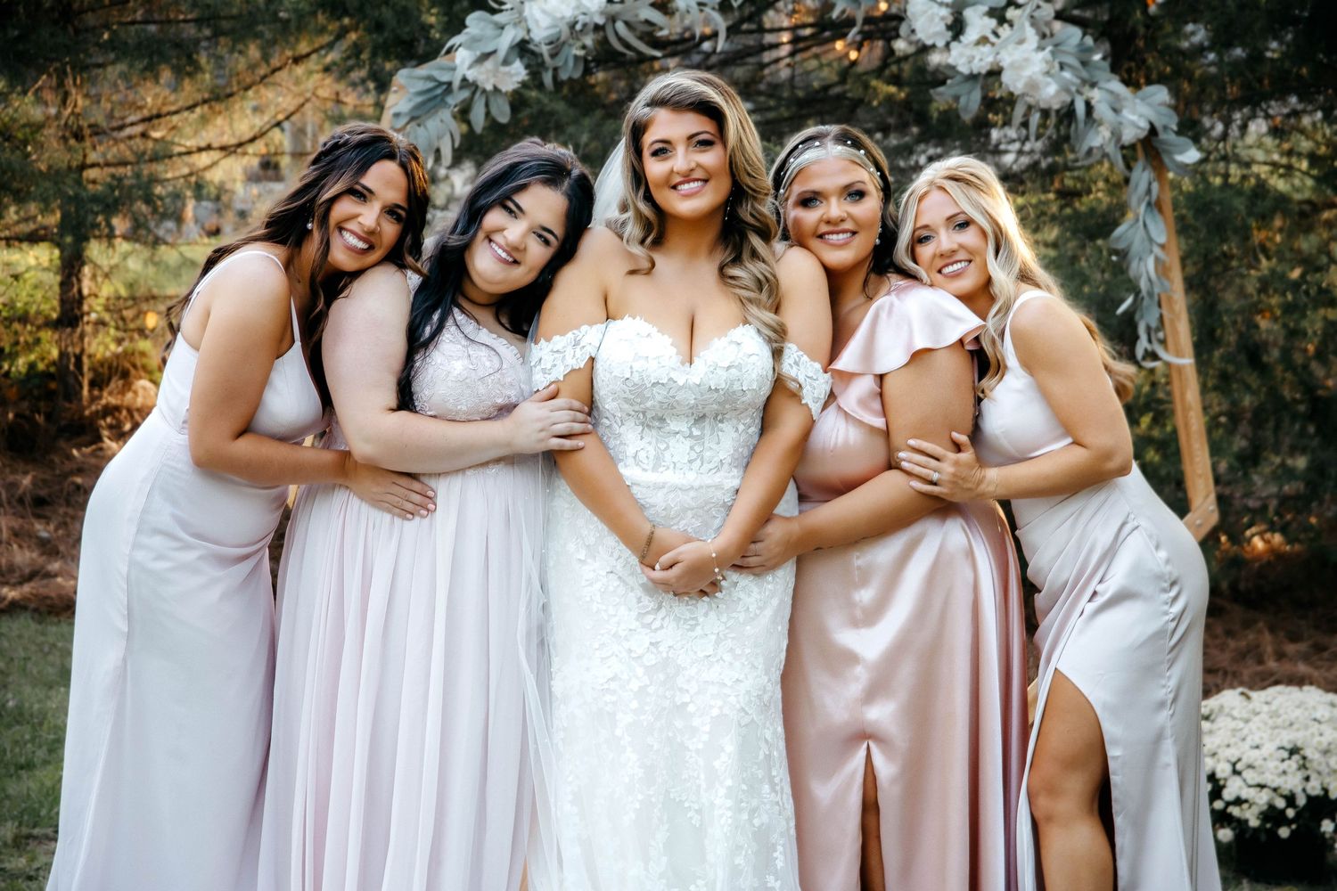 A group in light pink and white formal dresses pose together at an outdoor wedding celebration.
