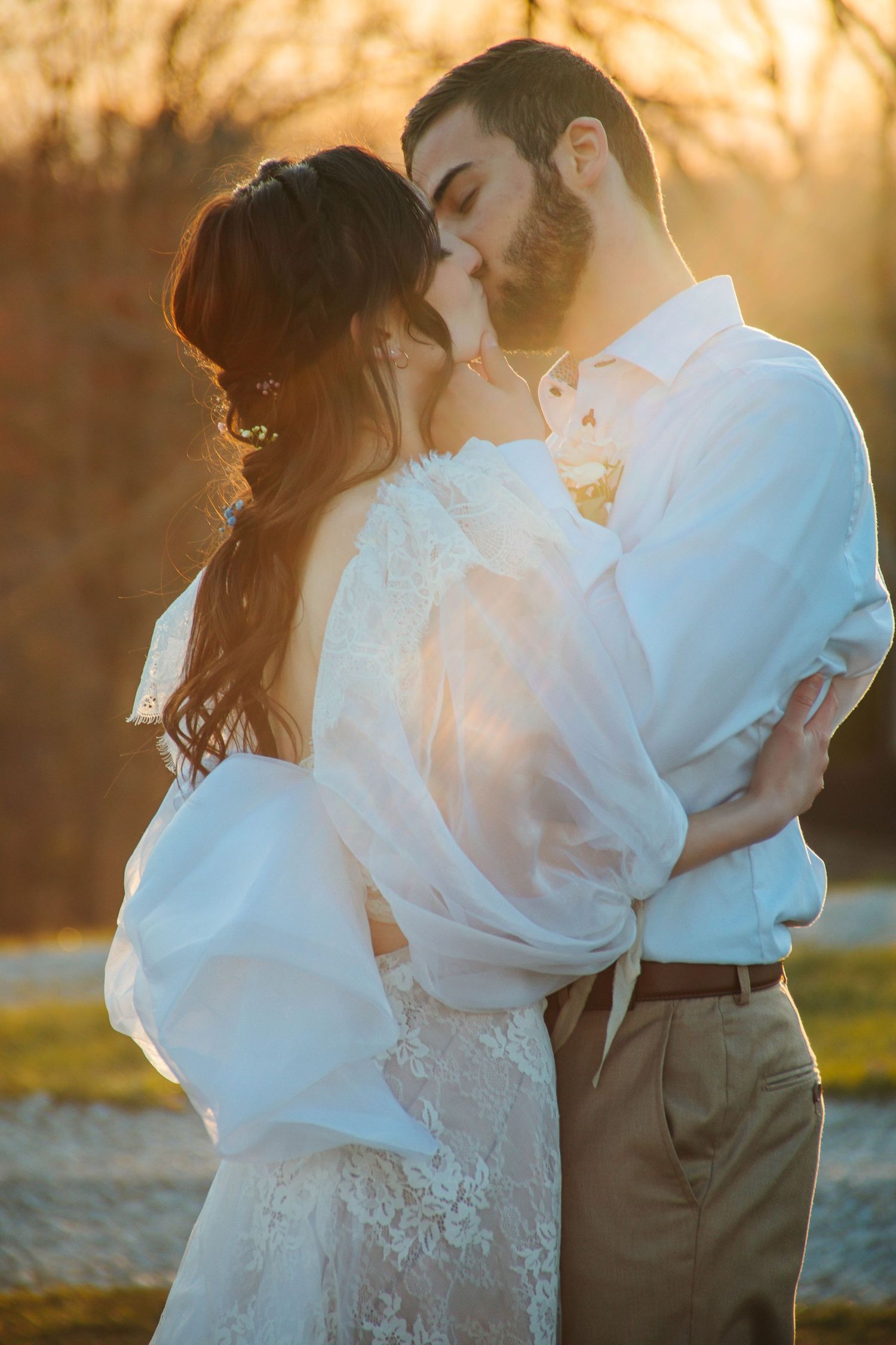 A romantic wedding couple embraces at sunset, the bride in a flowing white dress and groom in khaki pants and white shirt.