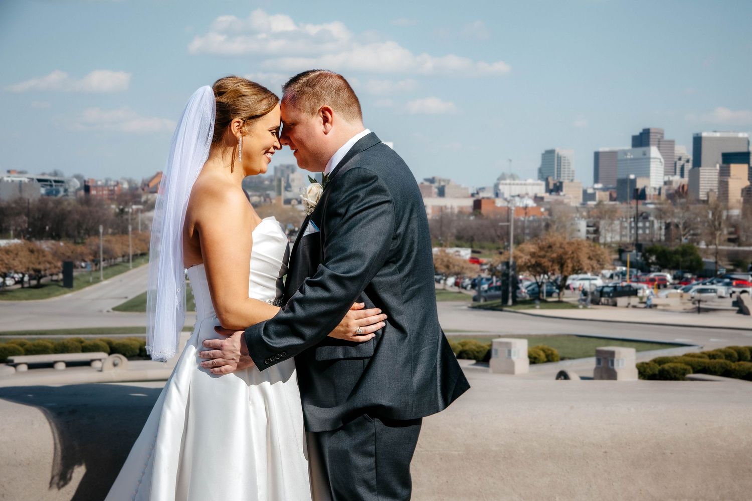 A couple in wedding attire embracing on a rooftop terrace with a city skyline in the background on a sunny day.