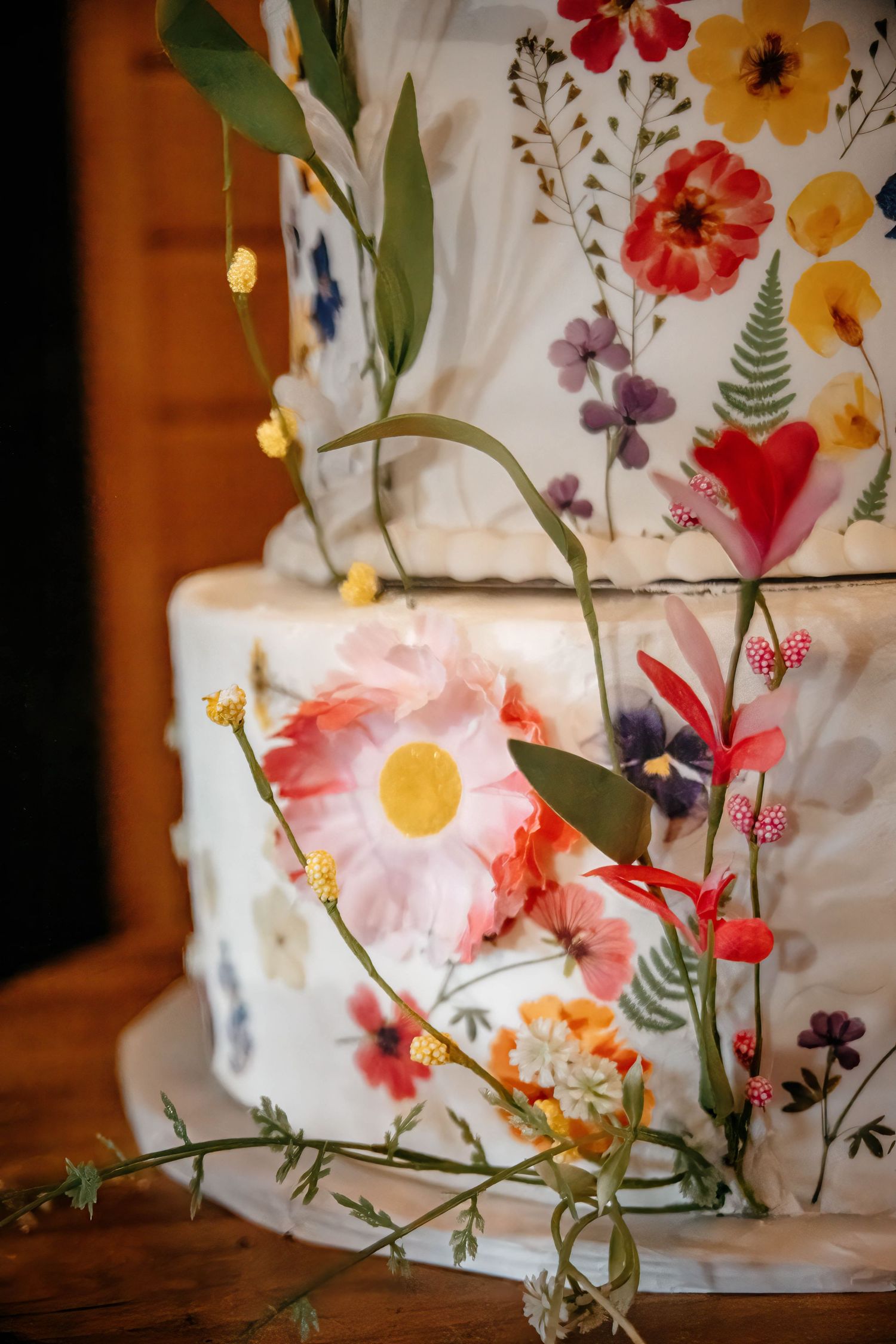 A white wedding cake decorated with pressed wildflowers and delicate floral designs against a warm wooden backdrop.