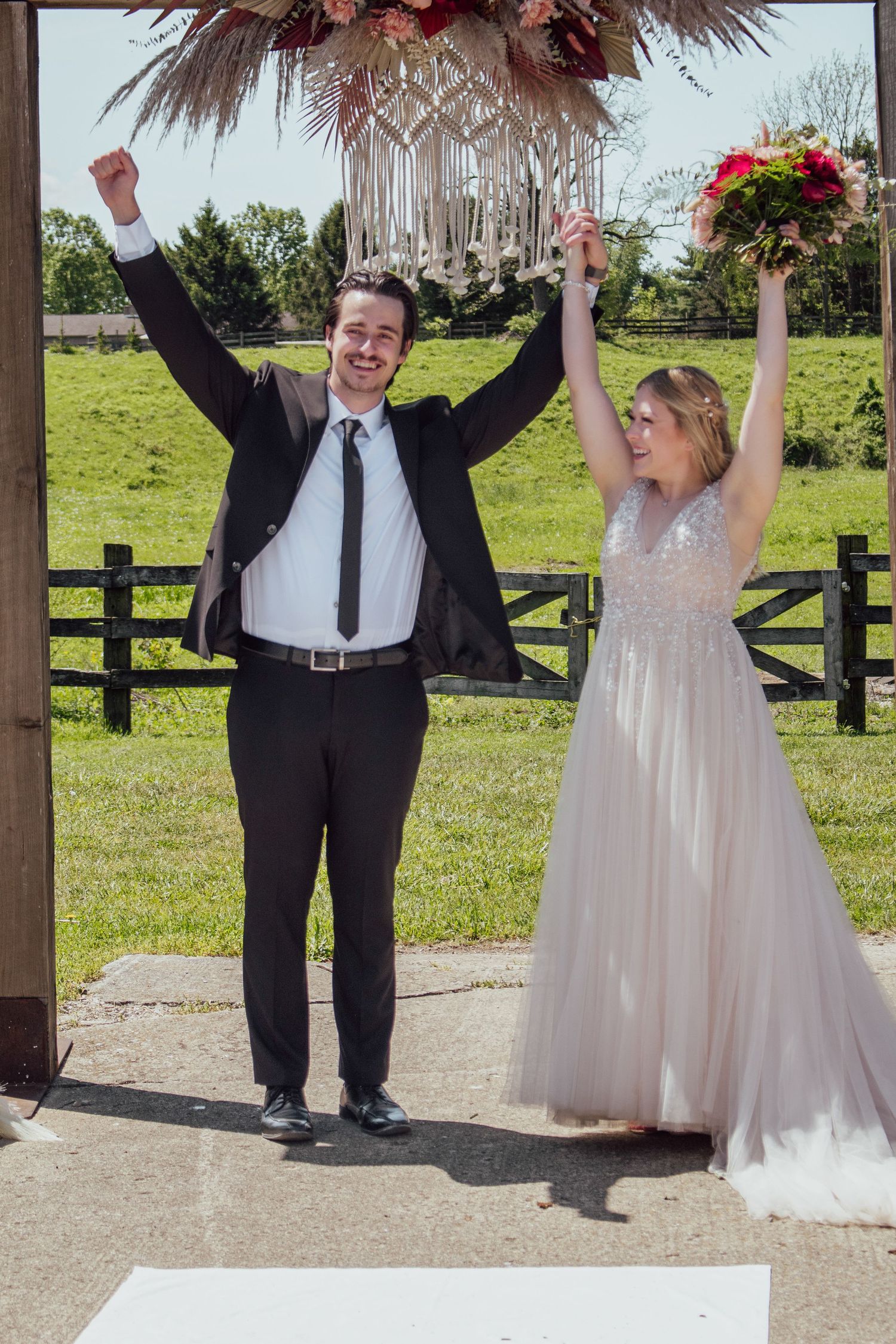 A newlywed couple celebrates with raised arms under a macrame arch on an outdoor patio with green fields in the background.