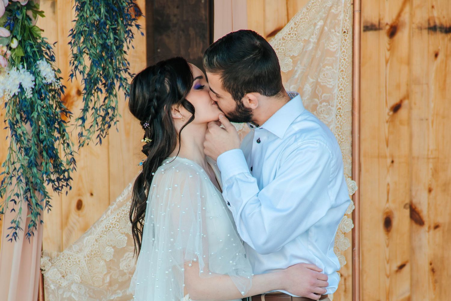 A romantic couple shares an intimate kiss against a rustic wooden wall decorated with hanging greenery and macrame.