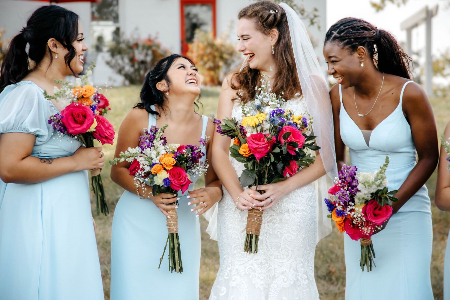 A bride and bridesmaids in light blue dresses share a joyful moment while holding vibrant pink and purple bouquets.