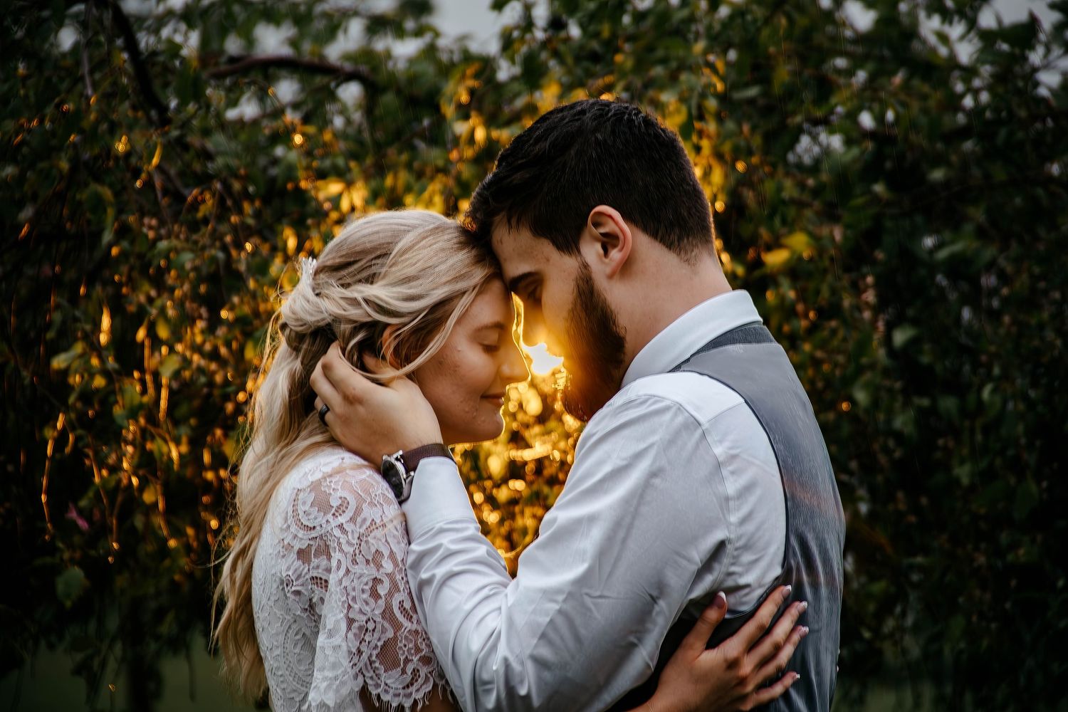 A romantic couple embraces in front of glowing sunset light through trees while dressed in wedding attire.